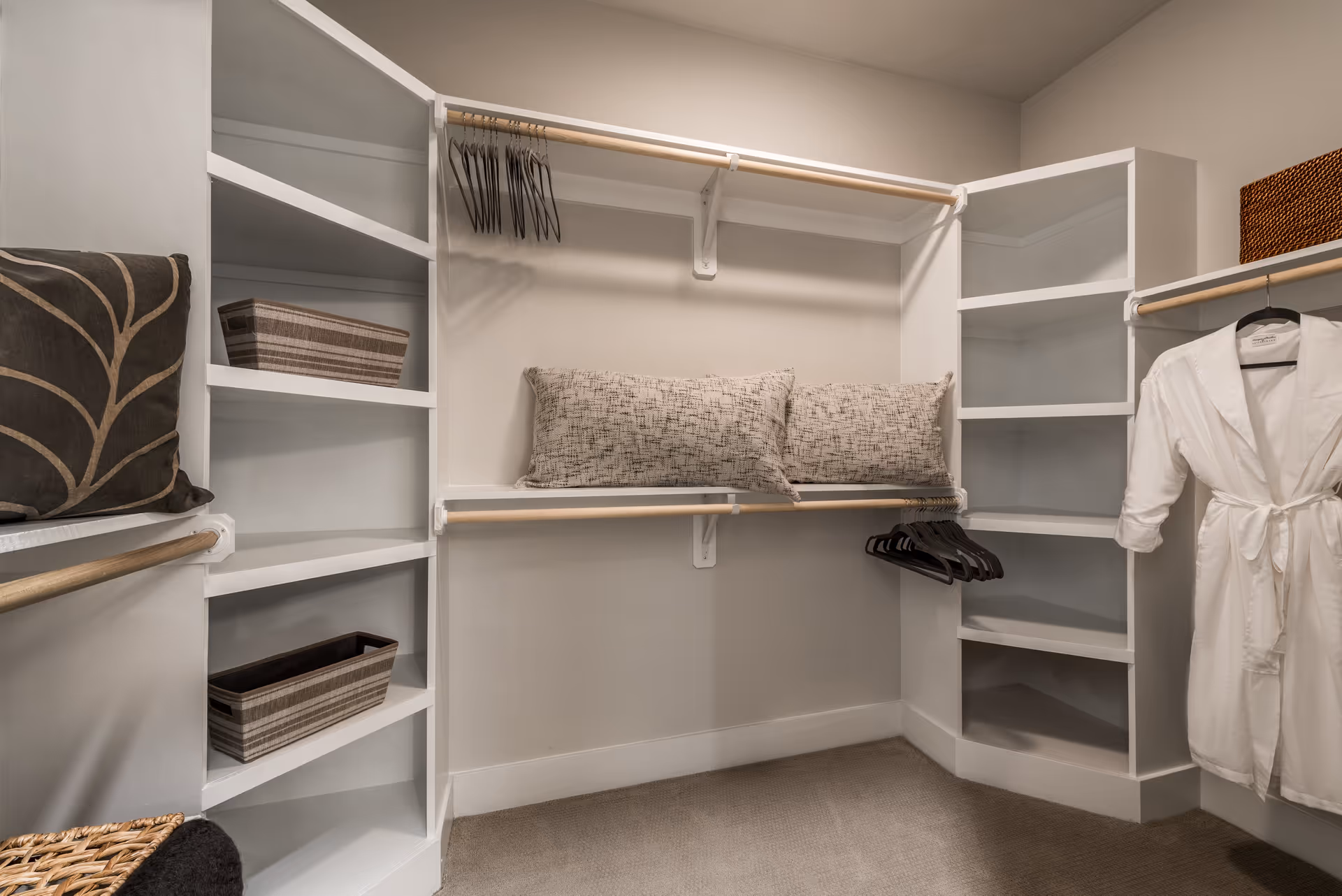 A spacious walk-in closet with white built-in shelves and hanging rods. The shelves hold decorative pillows and storage baskets, and several empty hangers are on the rods. A white robe is hanging on the right side.