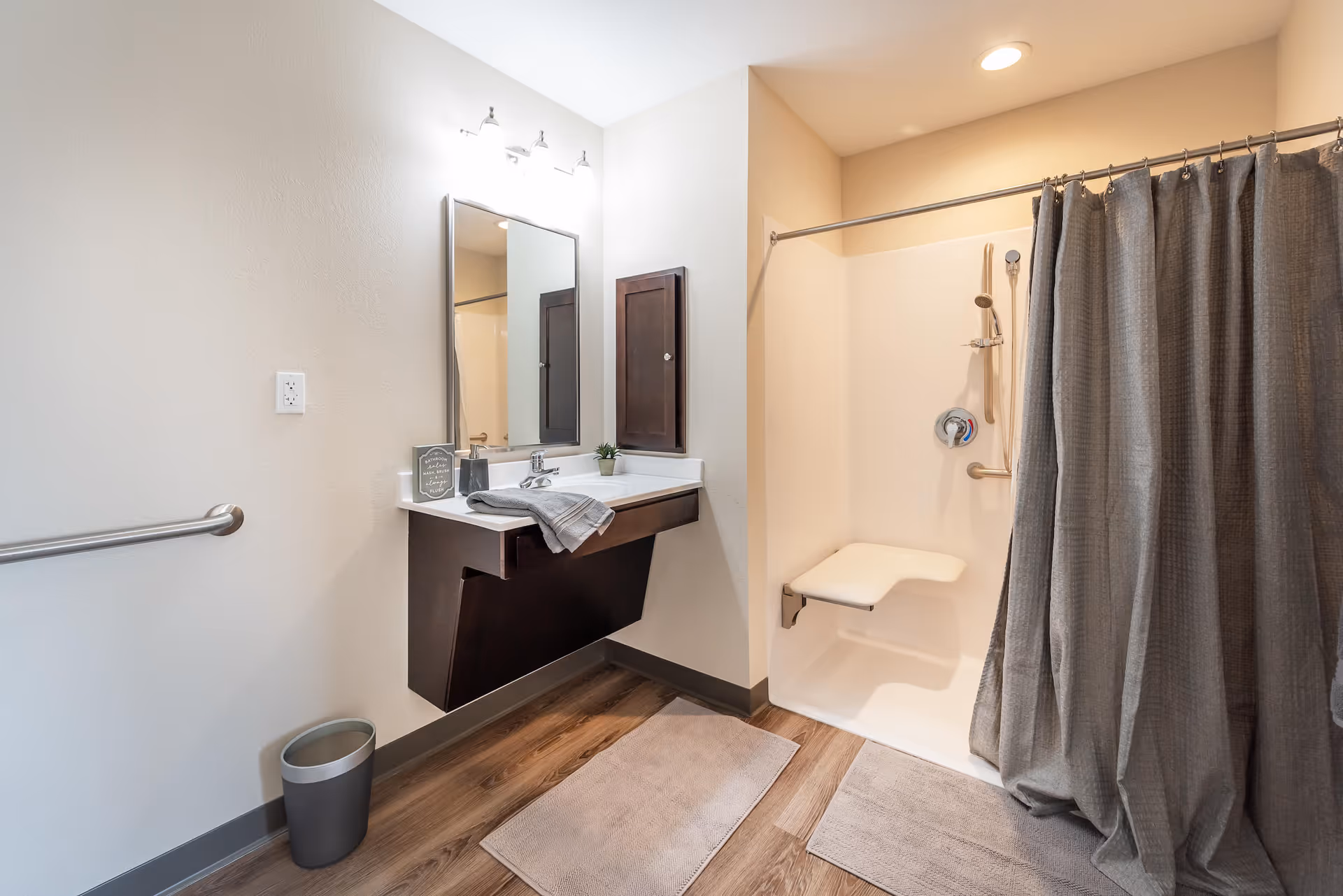 A clean, modern bathroom featuring a wall-mounted sink with a mirror and three-light fixture above it. There is a small potted plant and a folded towel on the sink. To the right, there is a walk-in shower with a fold-down seat and a gray shower curtain. The floor has wood-style flooring with two beige bath mats, and a small trash can is placed near the wall.