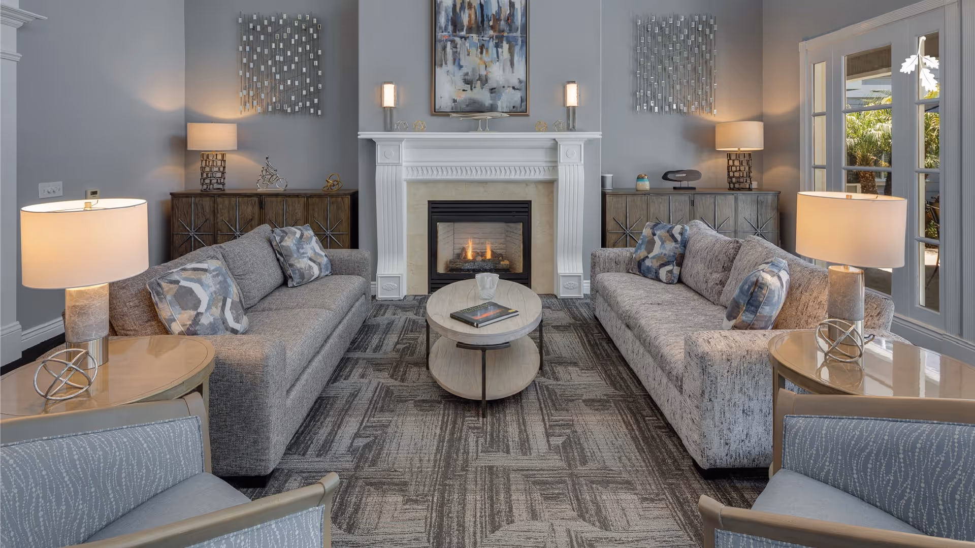 A cozy living room with two gray sofas facing a lit fireplace, a round coffee table, lamps, and artwork on the walls.
