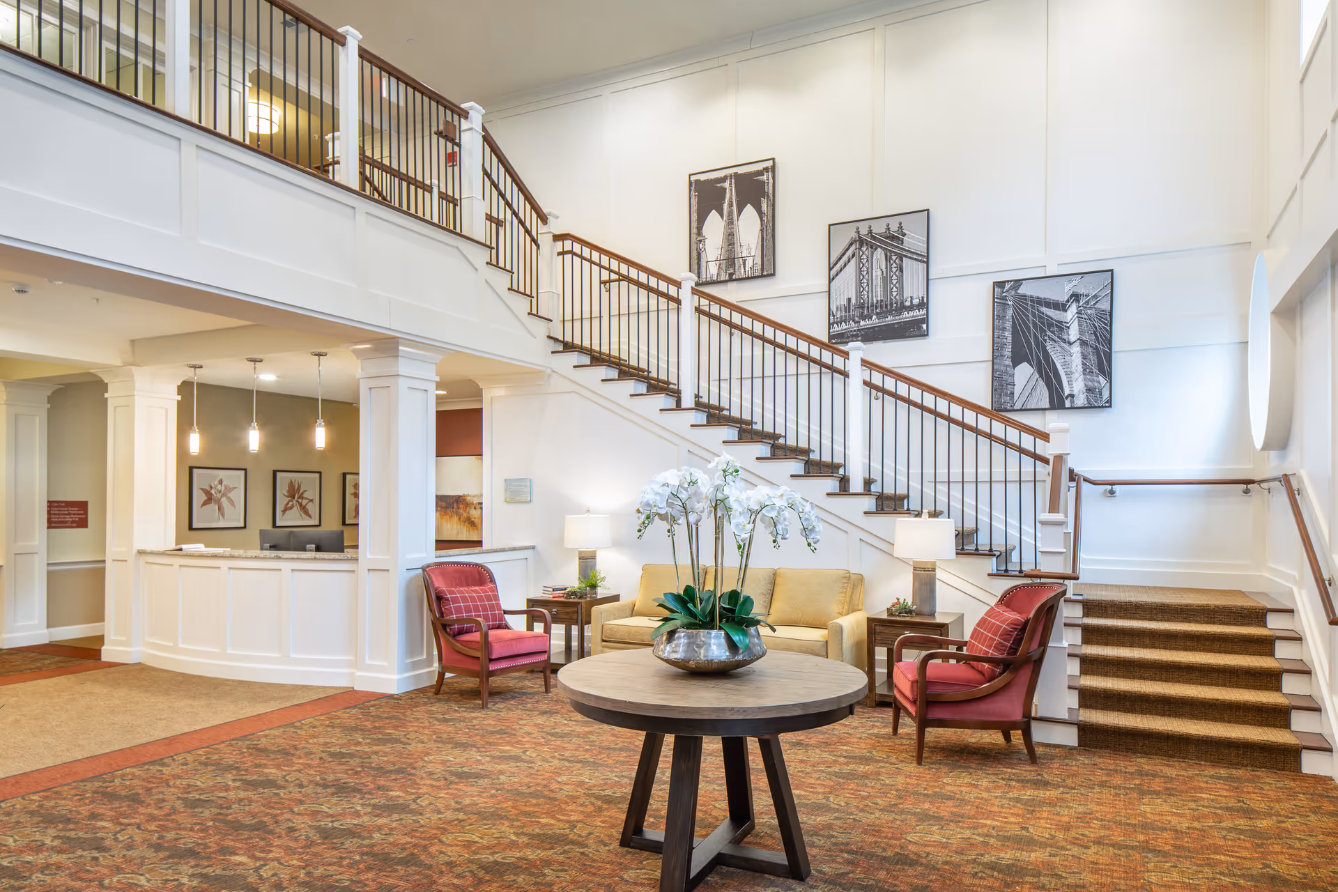 A bright and spacious senior living facility lobby with a staircase featuring wooden handrails and black balusters. The area includes a round wooden table with a silver vase holding white orchids, two red cushioned armchairs, a beige sofa, and side tables with lamps. The walls are decorated with black and white framed photographs of architectural structures. The reception desk is visible on the left side under hanging pendant lights.