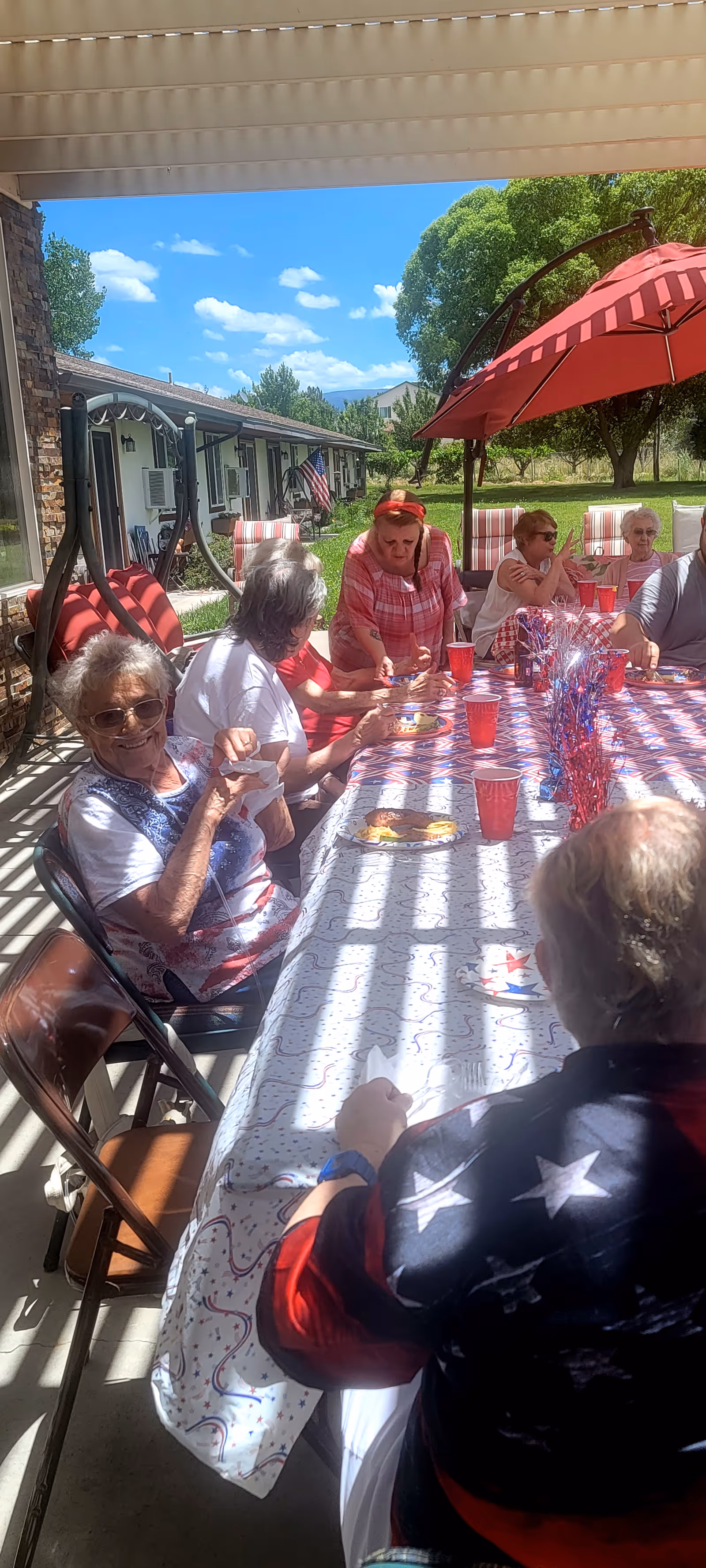 A group of elderly people sitting around a long table outdoors under a covered patio, enjoying a sunny day. The table is decorated with a festive tablecloth, red cups, and patriotic-themed decorations. There is a large red umbrella providing shade, and a building with an American flag is visible in the background along with green trees and a clear blue sky.