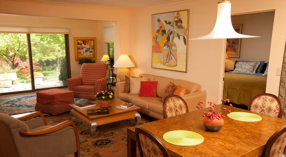A cozy living and dining area in a senior living facility with a beige sofa adorned with colorful pillows, a wooden coffee table with books and a flower pot, an armchair with an ottoman, and a dining table set with green placemats and a decorative centerpiece. A large window shows a view of a garden outside, and a bedroom with a bed and pillows is visible through an open doorway.