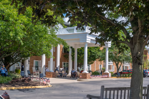 Covered entrance of a brick building with white columns, outdoor seating and people sitting under trees.