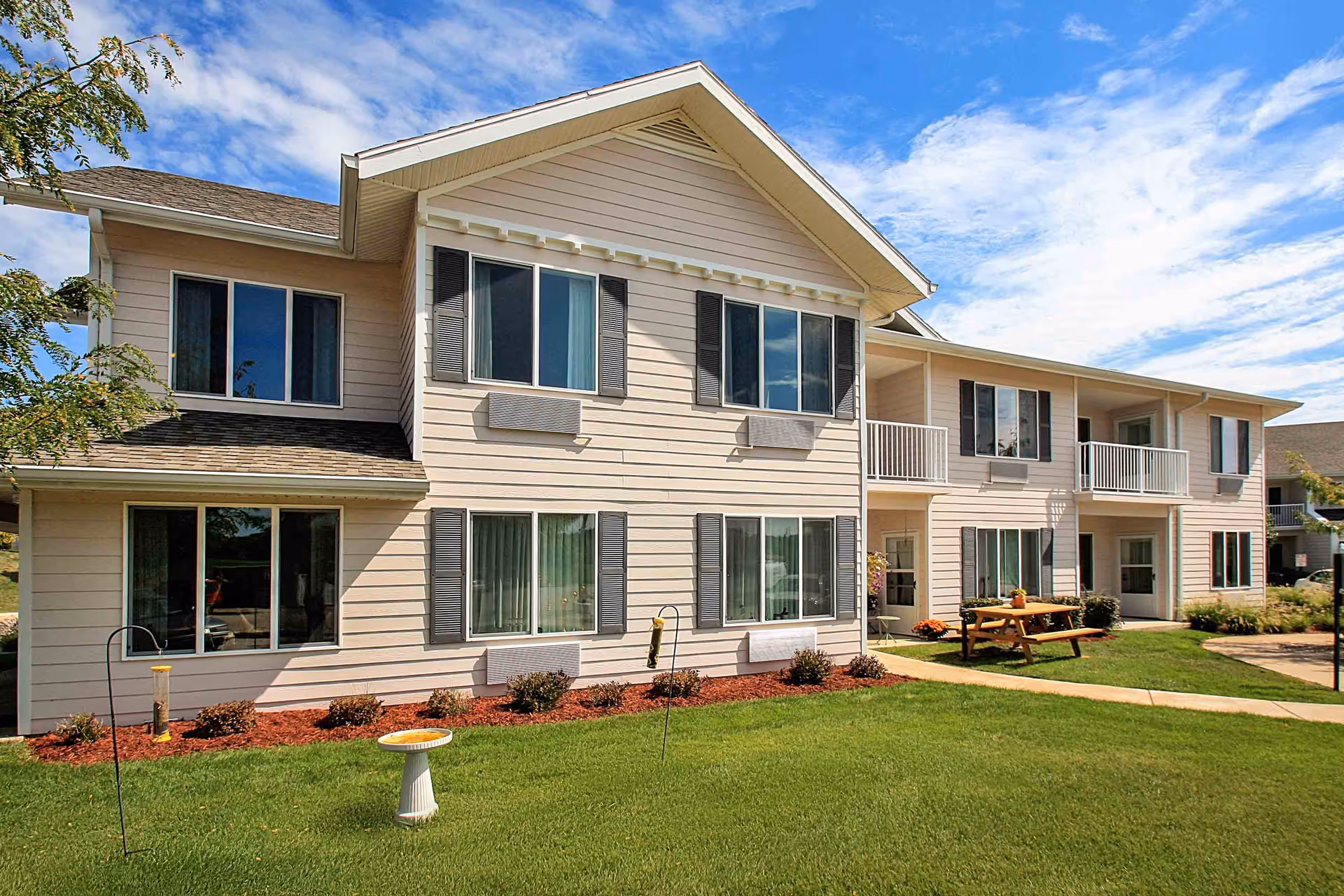 Exterior view of a two-story beige senior living facility building with multiple windows, small balconies, and a well-maintained lawn with a picnic table and bird feeders under a partly cloudy blue sky.