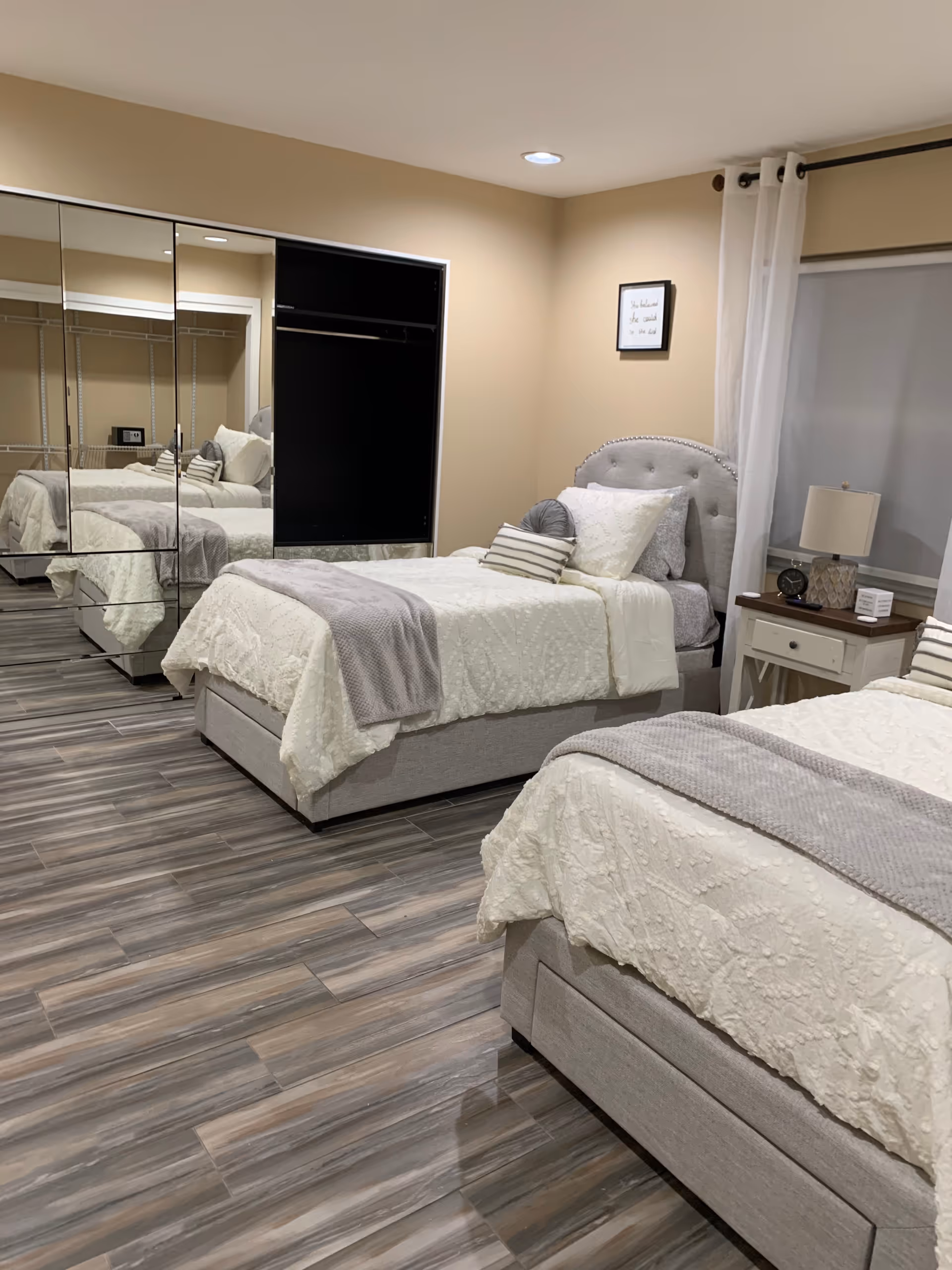 Twin beds with white bedding and gray accents in a neatly furnished bedroom with mirrored closet doors and wood-look flooring.