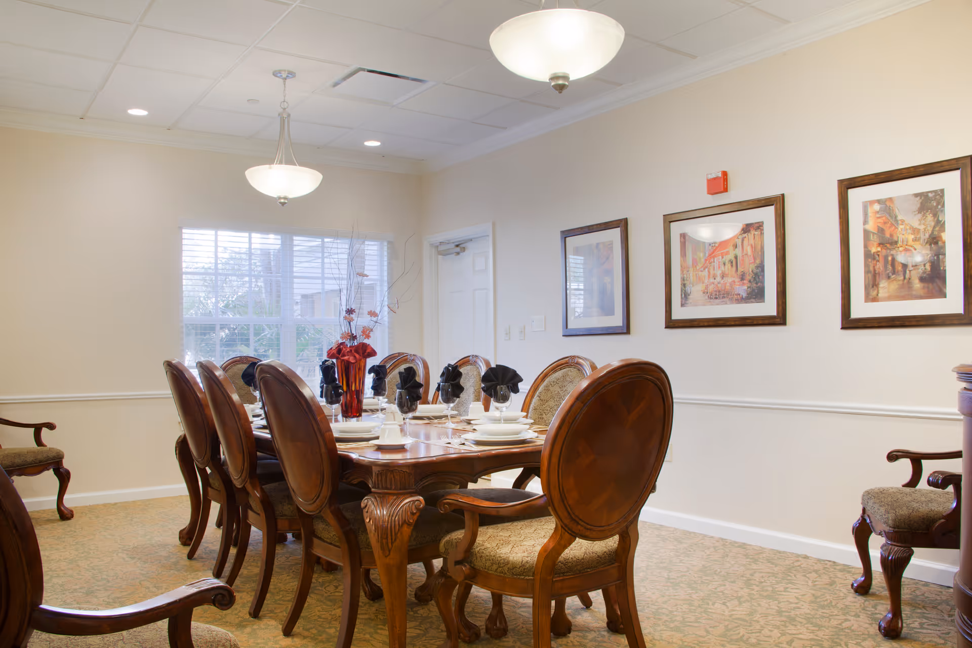 A formal dining room with a set wooden table and upholstered chairs, chandeliers, place settings, and framed artwork on the walls.
