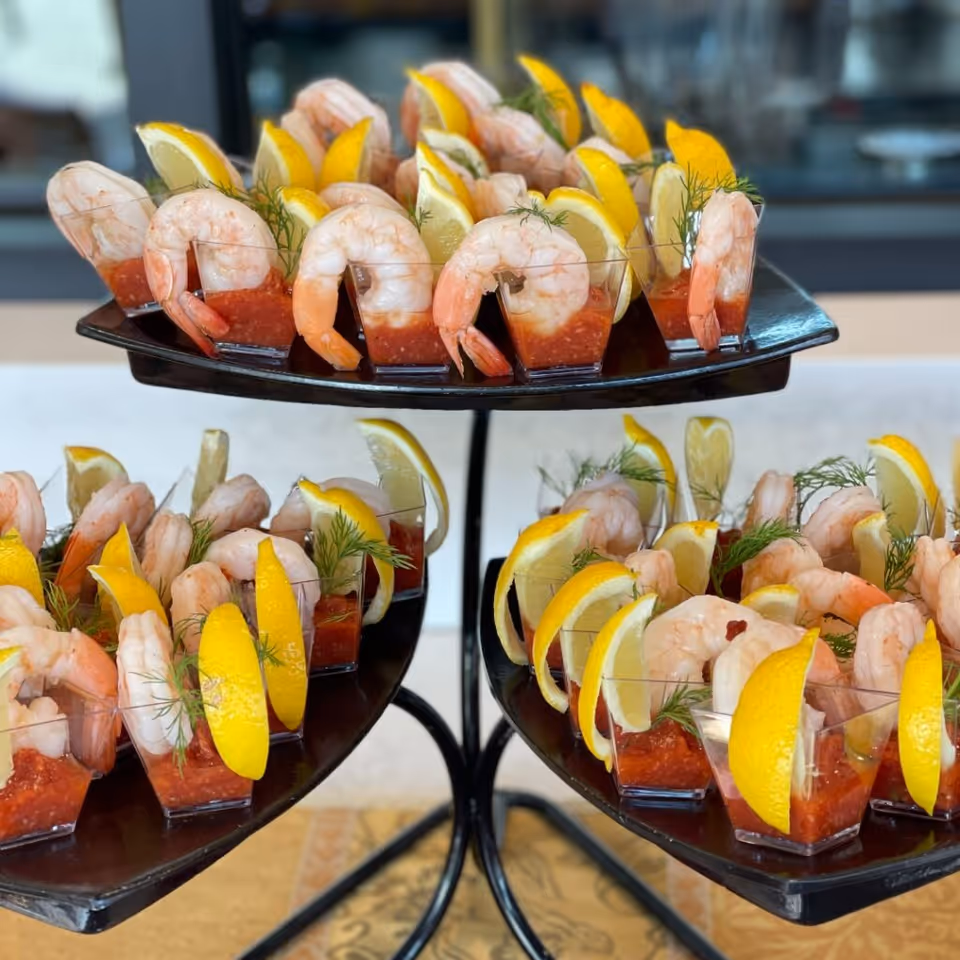 Three-tiered serving tray with individual servings of shrimp cocktail, each with a shrimp, cocktail sauce, a lemon wedge, and a sprig of dill in a small clear cup.