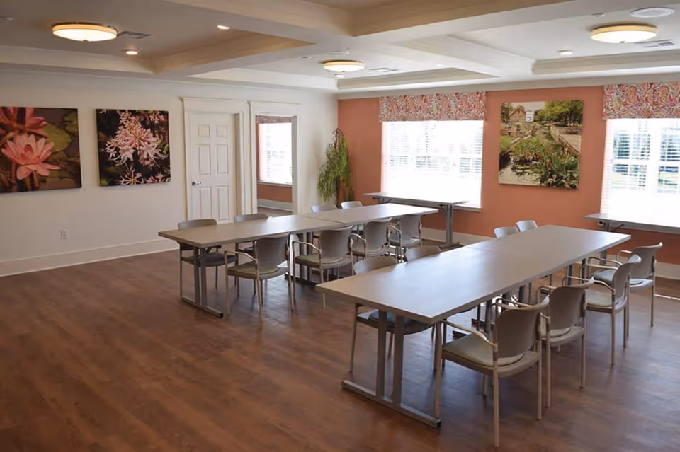 A bright room with wooden flooring featuring two long tables arranged parallel with multiple chairs around them. The walls are decorated with floral and garden-themed paintings, and there are large windows with blinds letting in natural light. The ceiling has recessed lighting fixtures.