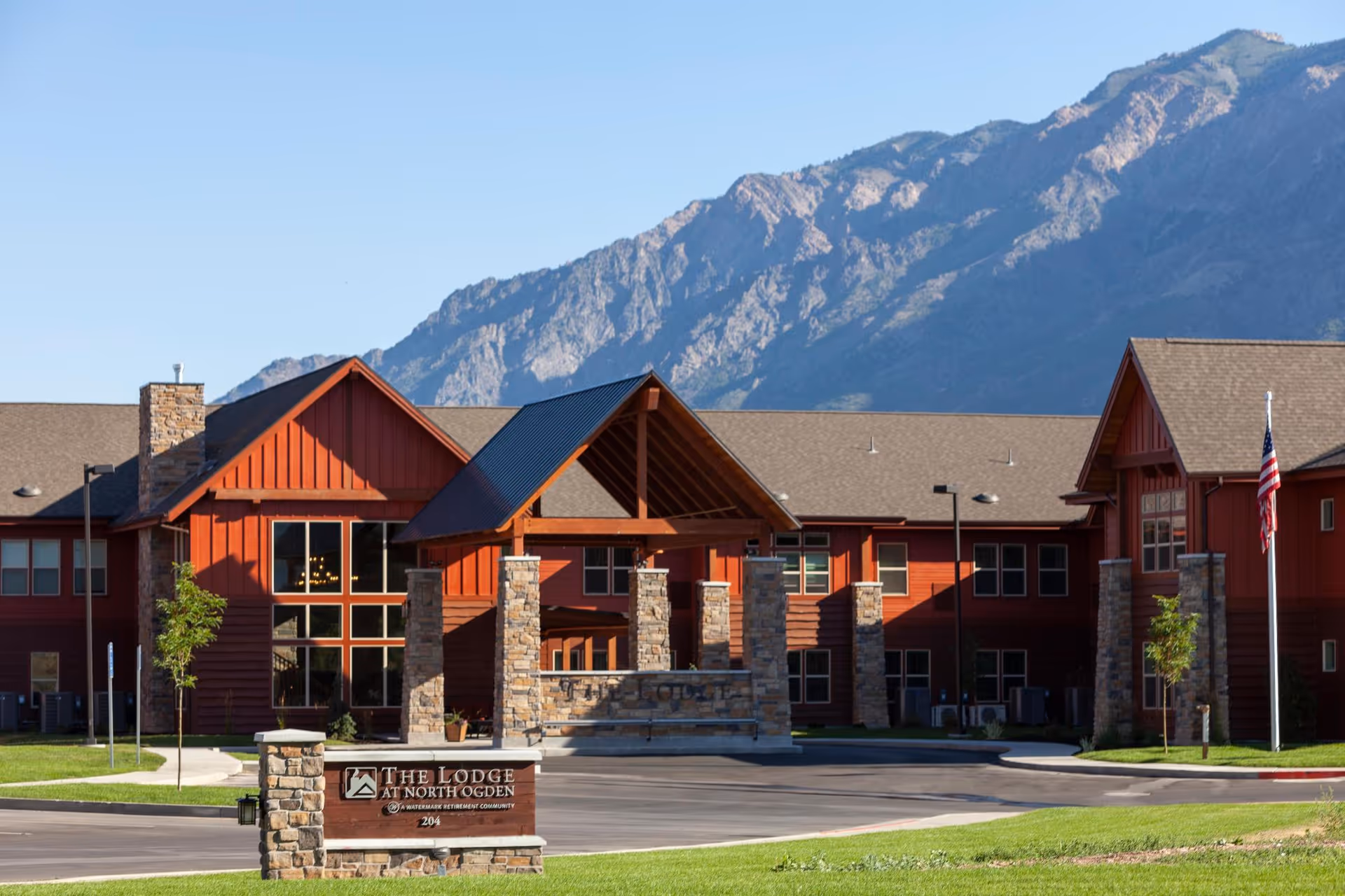 Front exterior of The Lodge at North Ogden senior living building with stone entrance and mountains in the background.