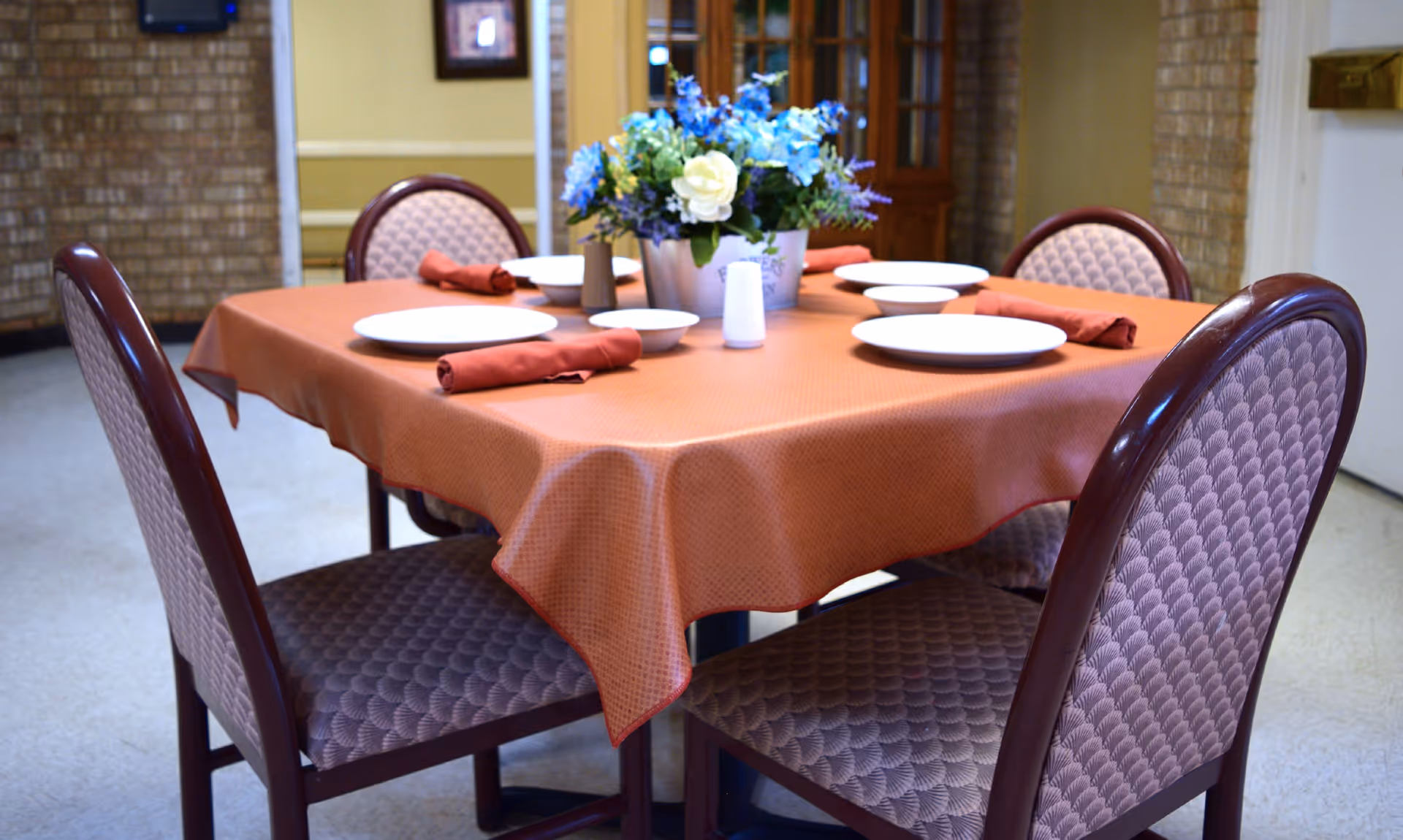 A dining table set for four with a rust-colored tablecloth, white plates, bowls, and rolled red napkins. A floral centerpiece with blue and white flowers is in the middle of the table. Four upholstered chairs with patterned fabric surround the table. The background shows a brick wall and a wooden cabinet.
