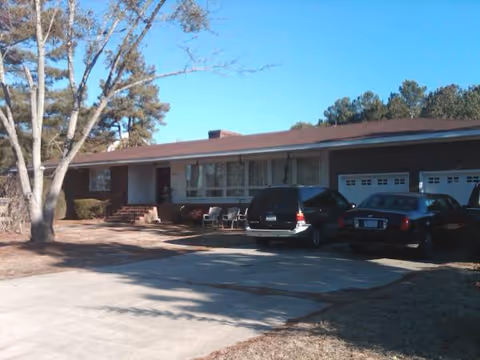 Single-story residential-style senior home exterior with driveway, parked cars, and front entrance steps.