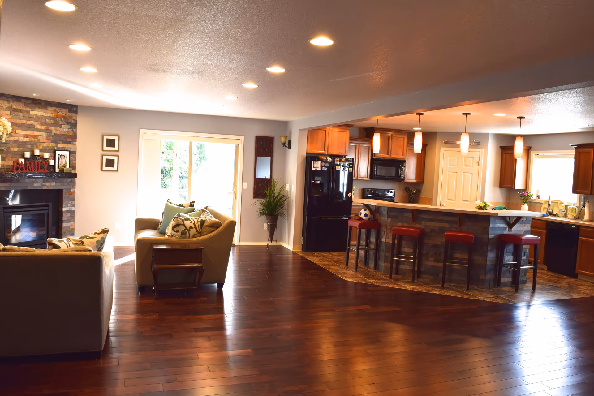 Open-plan living room and kitchen with hardwood floors, a stone fireplace, seating area, and a breakfast bar with stools.
