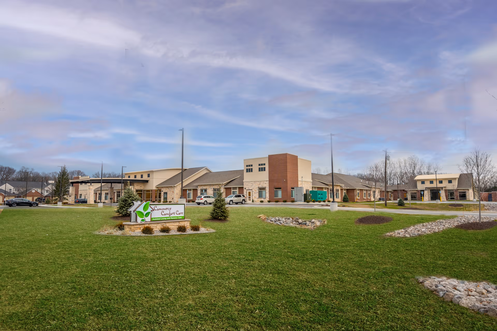 Wide exterior view of a senior living facility building with a large green lawn in the foreground. The building has a modern design with multiple sections, including brick and stone facades. There is a sign on the lawn that reads 'Commerce Comfort Care Advanced Living and Rehab Center.' Several cars are parked near the building under a partly cloudy sky.