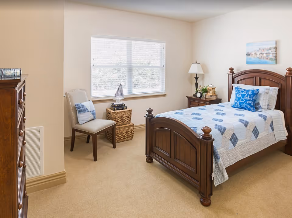 A cozy bedroom with a wooden bed frame and a blue and white quilted bedspread. There is a matching blue pillow with sailboat designs on the bed. Next to the bed is a wooden nightstand with a lamp, a clock, and decorative items. A window with white blinds lets in natural light. A beige chair with a blue and white pillow sits beside two woven baskets stacked on top of each other. A wooden dresser is partially visible on the left side of the image. The walls are painted light beige and a framed picture hangs above the bed.
