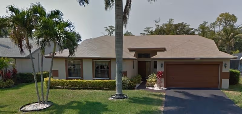 Single-story residential building with a brown shingled roof, beige walls, and a two-car garage. The front yard has green grass, palm trees, and small bushes along the house. The driveway is paved and leads to the garage.