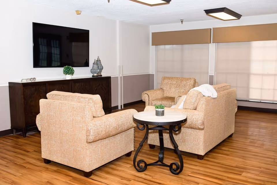 A cozy living room area with three beige upholstered armchairs arranged around a small round table with a potted plant. A large flat-screen TV is mounted on the wall above a dark wooden cabinet. The room has wooden flooring and large windows with beige blinds.