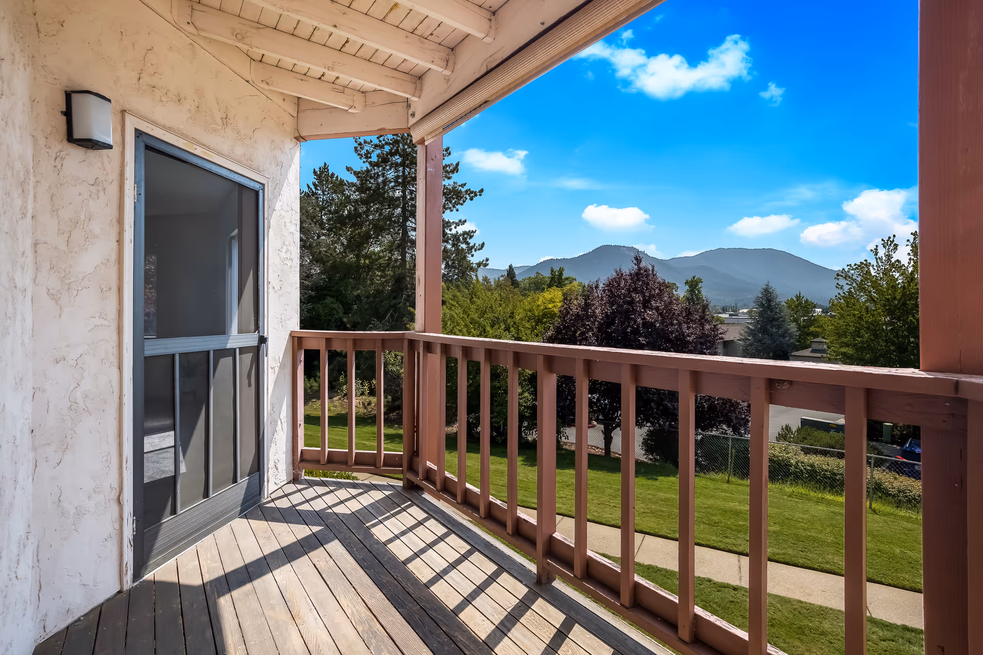 Wooden balcony with a railing overlooking landscaped grounds and distant mountains under a blue sky.