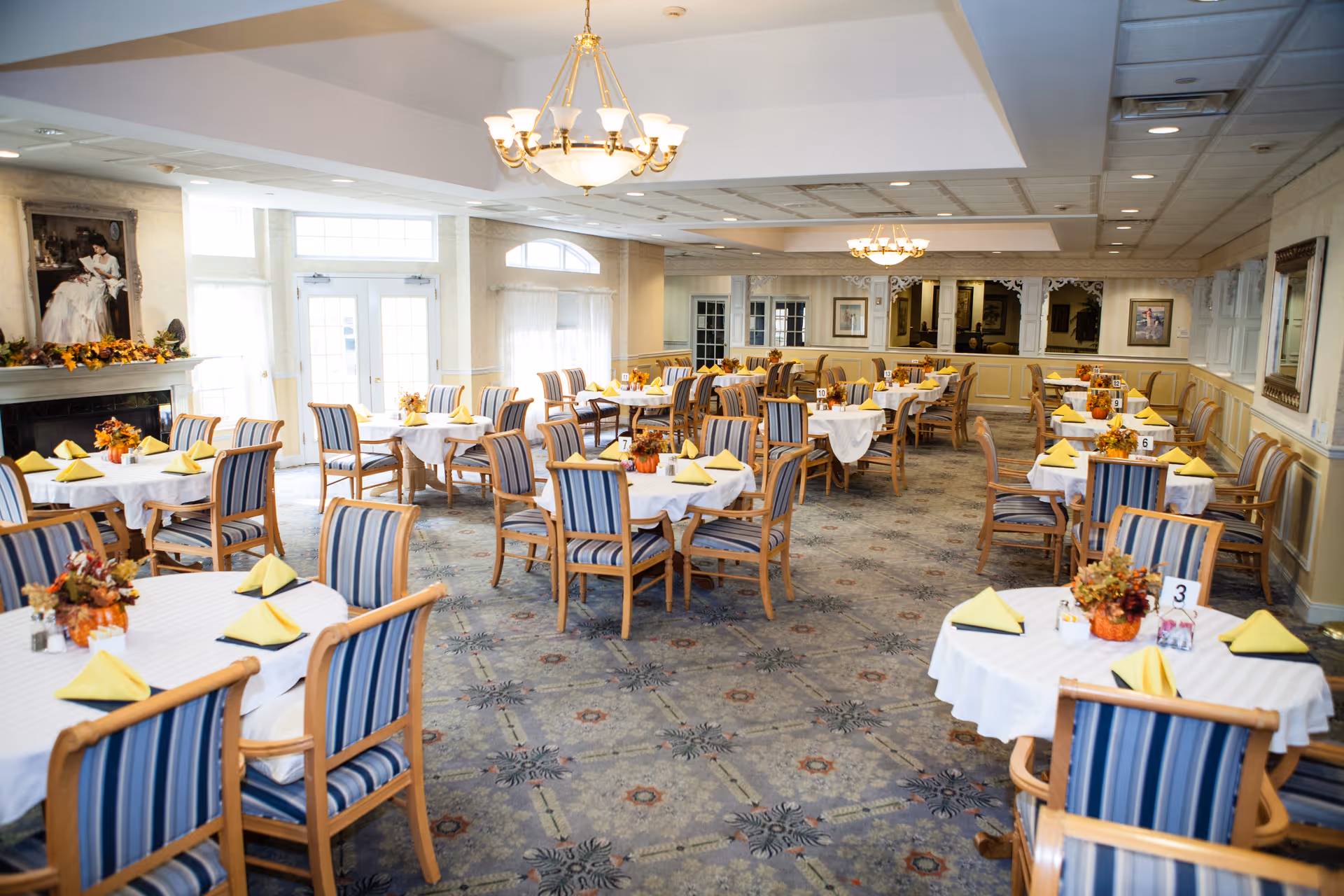 A spacious dining room with multiple round tables covered in white tablecloths, each set with yellow folded napkins and autumn-themed floral centerpieces. The room features striped wooden chairs, chandeliers hanging from the ceiling, large windows letting in natural light, and a fireplace with a painting above it.