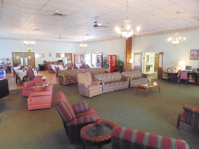 A spacious common area in a senior living facility featuring multiple seating arrangements with patterned sofas and armchairs, a coffee table with flowers, a dining area with tables and chairs, a Coca-Cola vending machine, and a desk with a computer and lamp against the wall.