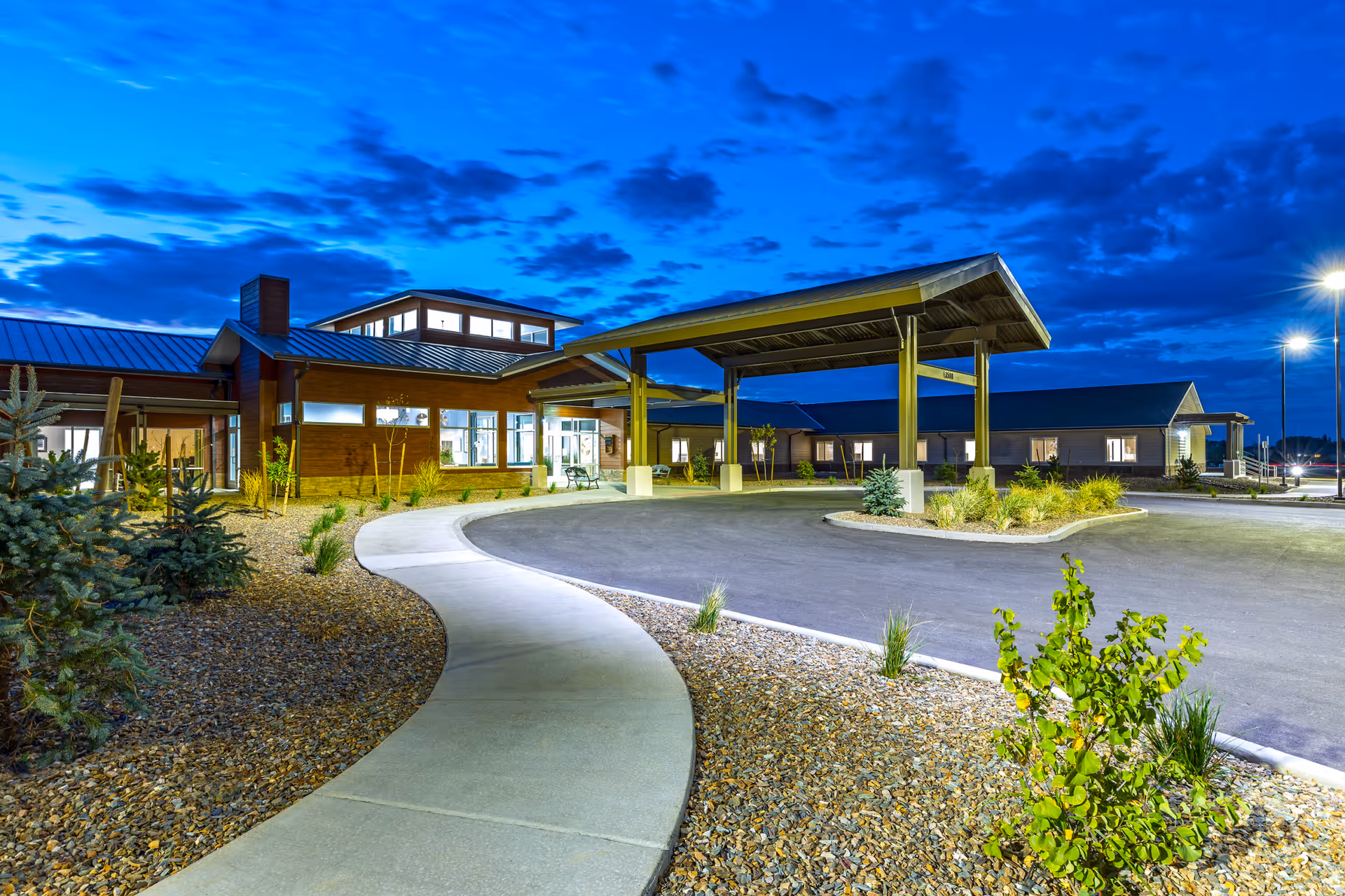 Exterior view of The Landings Of Prescott Valley facility at dusk, showing a curved sidewalk leading to a covered entrance with well-lit windows and landscaped surroundings including small trees and shrubs under a dramatic blue sky with scattered clouds.