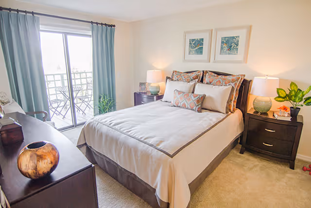 Bright bedroom with a neatly made bed, two nightstands with lamps, a dresser, and a sliding glass door leading to a small balcony.