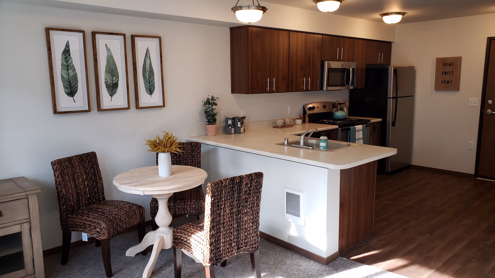 A cozy kitchen and dining area featuring a small round wooden table with a white vase holding yellow flowers, surrounded by three woven chairs. The kitchen has dark wood cabinets, a stainless steel microwave, stove, and refrigerator. On the wall above the table are three framed botanical prints of green leaves. A sign on the far wall reads 'HOME SWEET HOME'.