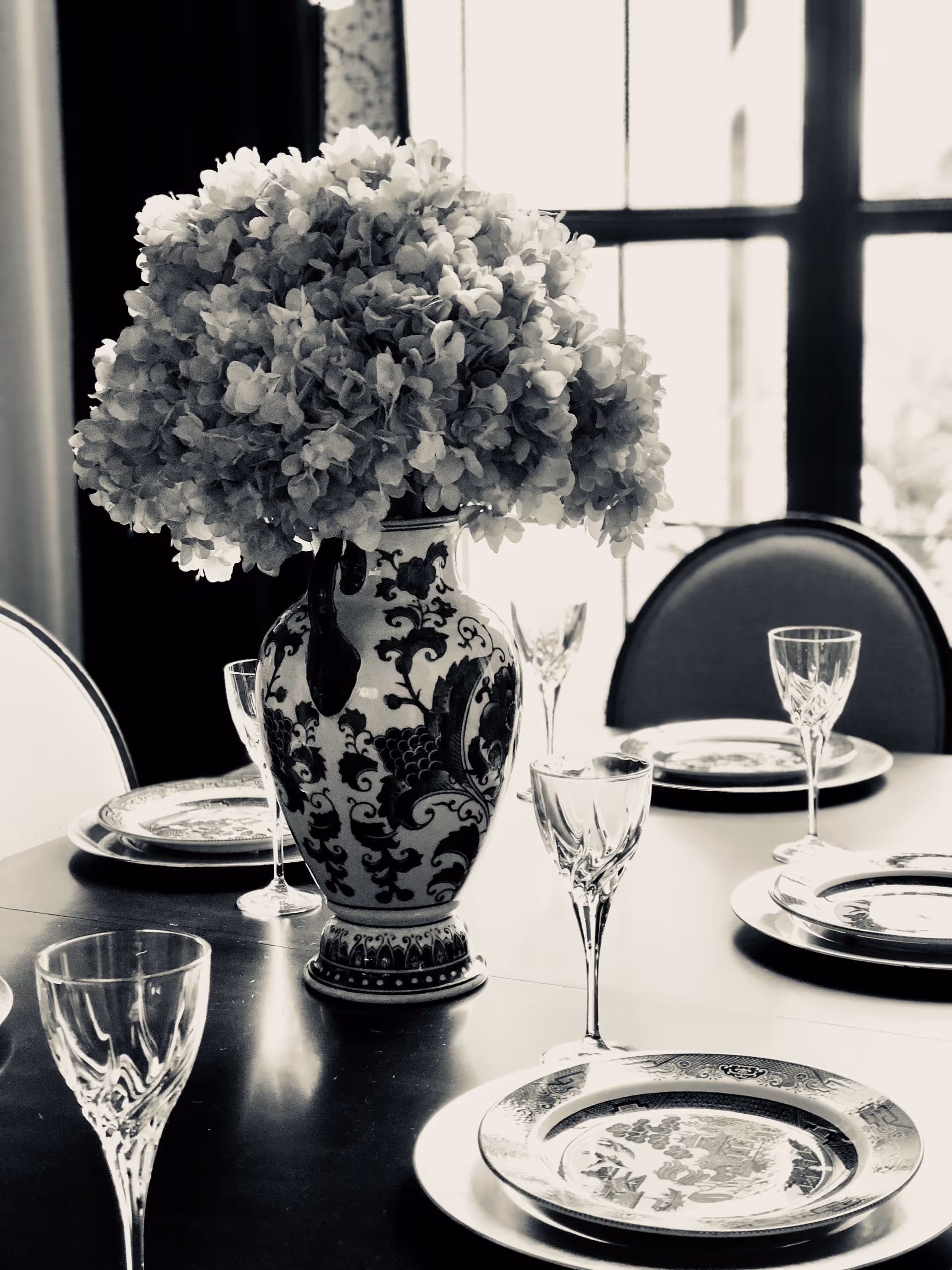 A dining table set with decorative plates and crystal wine glasses, featuring a large ornate vase filled with a bouquet of flowers as the centerpiece, with a window in the background.