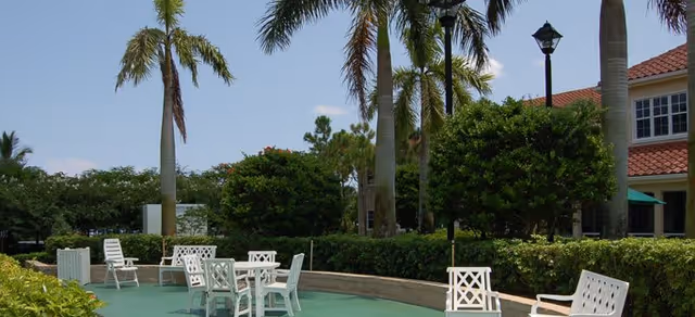 Outdoor seating area with white chairs and tables on a green surface surrounded by palm trees and bushes, with a building and lamppost in the background under a partly cloudy sky.