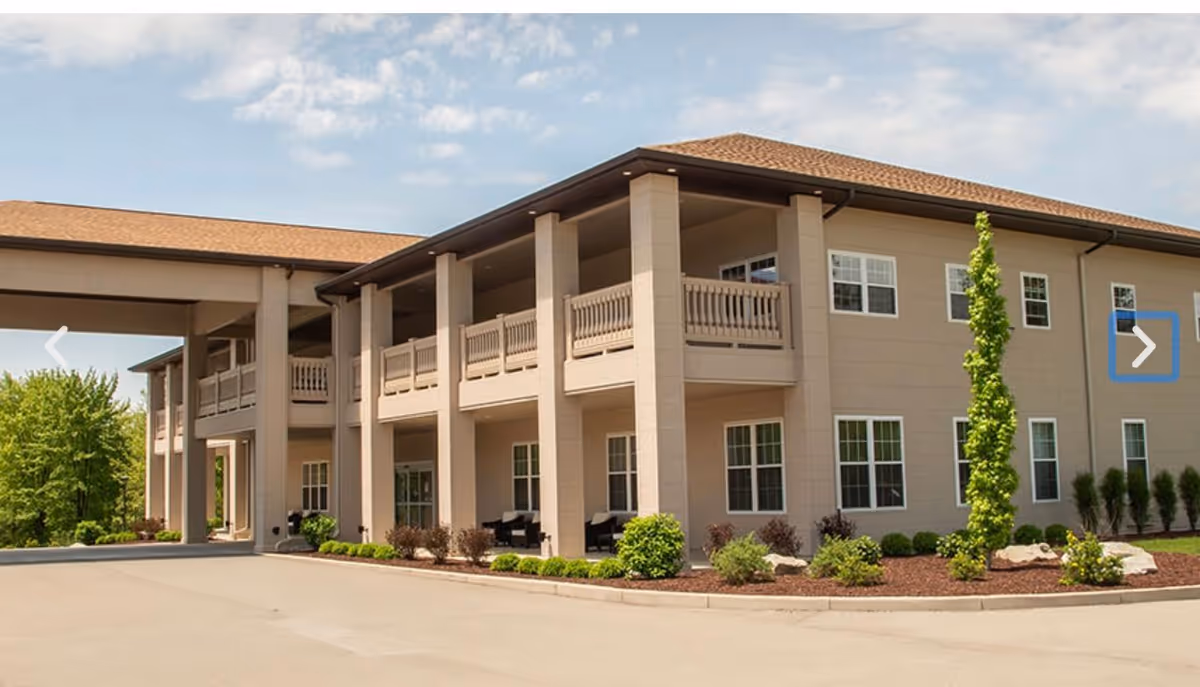 Exterior view of a two-story senior living facility building with beige walls, multiple windows, and a covered entrance. The building is surrounded by landscaped bushes, small trees, and a paved driveway under a partly cloudy sky.