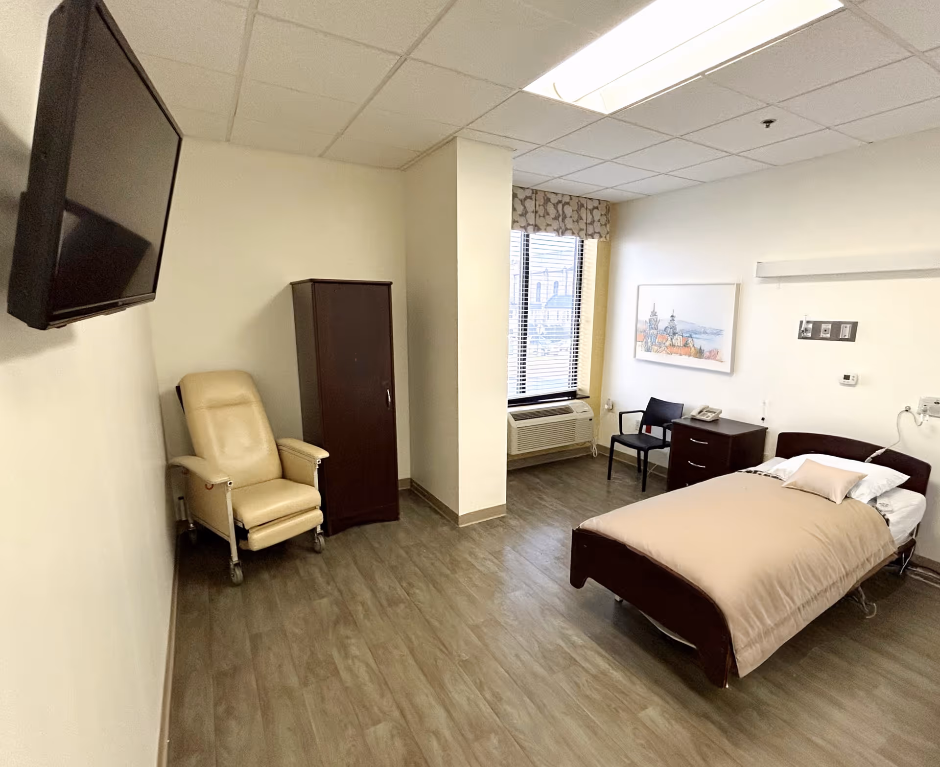 A simple, clean bedroom in a senior living facility with a single bed covered in beige bedding, a small nightstand with a telephone, a black chair, a window with blinds and a patterned valance, a wall-mounted flat screen TV, a beige recliner chair, and a dark wooden wardrobe. The floor is wood-style laminate and the walls are light-colored.