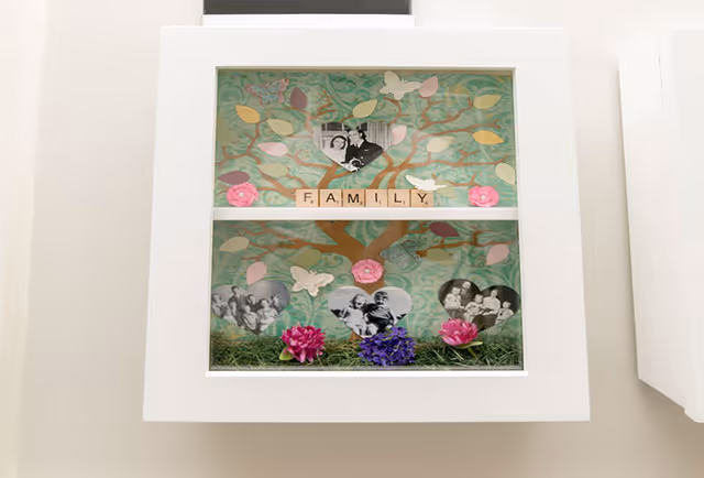 A white-framed shadow box featuring a decorative family tree with pastel-colored leaves, pink and purple flowers, and black-and-white family photos in heart shapes. The word FAMILY is spelled out in Scrabble tiles across the middle.