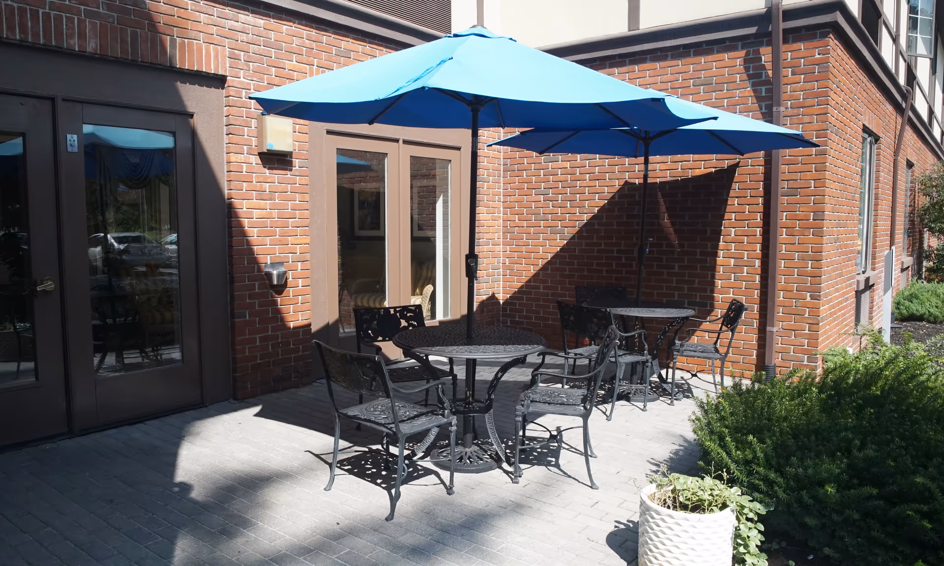 Brick-walled outdoor patio with black metal tables and chairs shaded by two blue umbrellas and a potted plant.