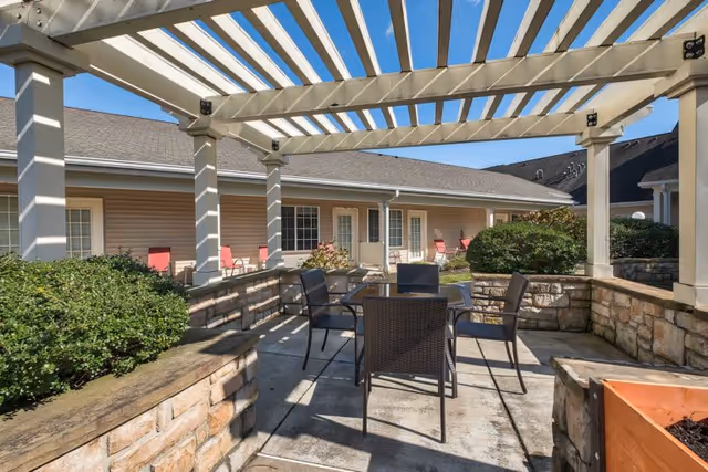 Outdoor patio area with a pergola overhead, featuring a glass-top table surrounded by four black chairs. The patio is enclosed by low stone walls with greenery and bushes around. In the background, there is a single-story building with beige siding, white trim, and several doors and windows. The sky is clear and blue.