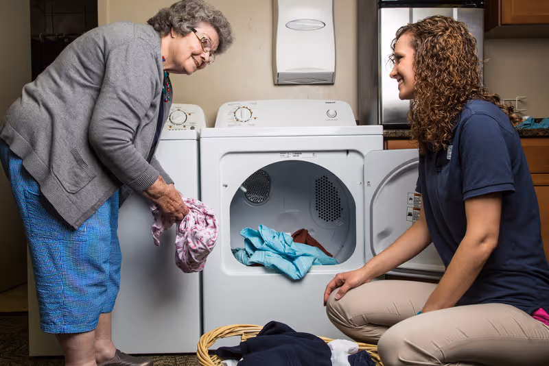 An elderly woman and a younger woman are doing laundry together in a laundry room. The elderly woman is standing and holding a piece of clothing, while the younger woman is kneeling in front of an open dryer with clothes inside. There is a washing machine next to the dryer and cabinets above a countertop in the background.