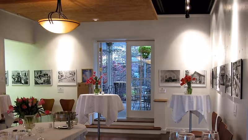 Interior view of a well-lit room with white walls adorned with black and white photographs. The room contains several tall round tables covered with white tablecloths and decorated with vases of pink and red flowers. Through glass doors at the back, an outdoor patio area with chairs and greenery is visible.
