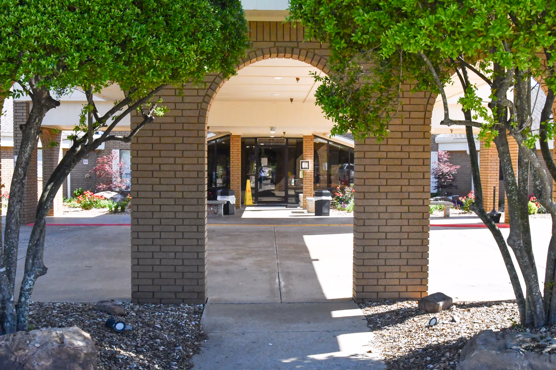 View of the entrance to Oaks Healthcare Center showing a covered walkway supported by brick columns with trees and landscaping on either side.