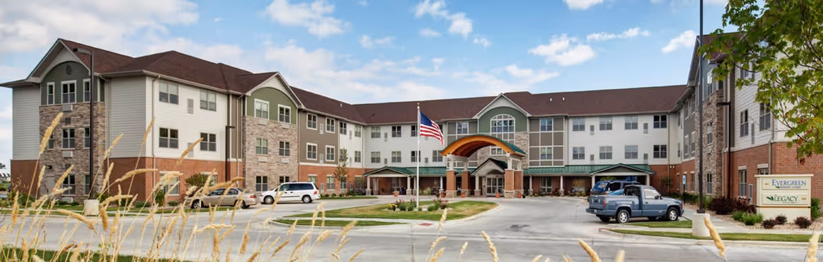 Front exterior view of a large three-story senior living facility building with a covered entrance, multiple windows, an American flag on a flagpole, several parked vehicles, and landscaping including grass and trees under a partly cloudy sky.