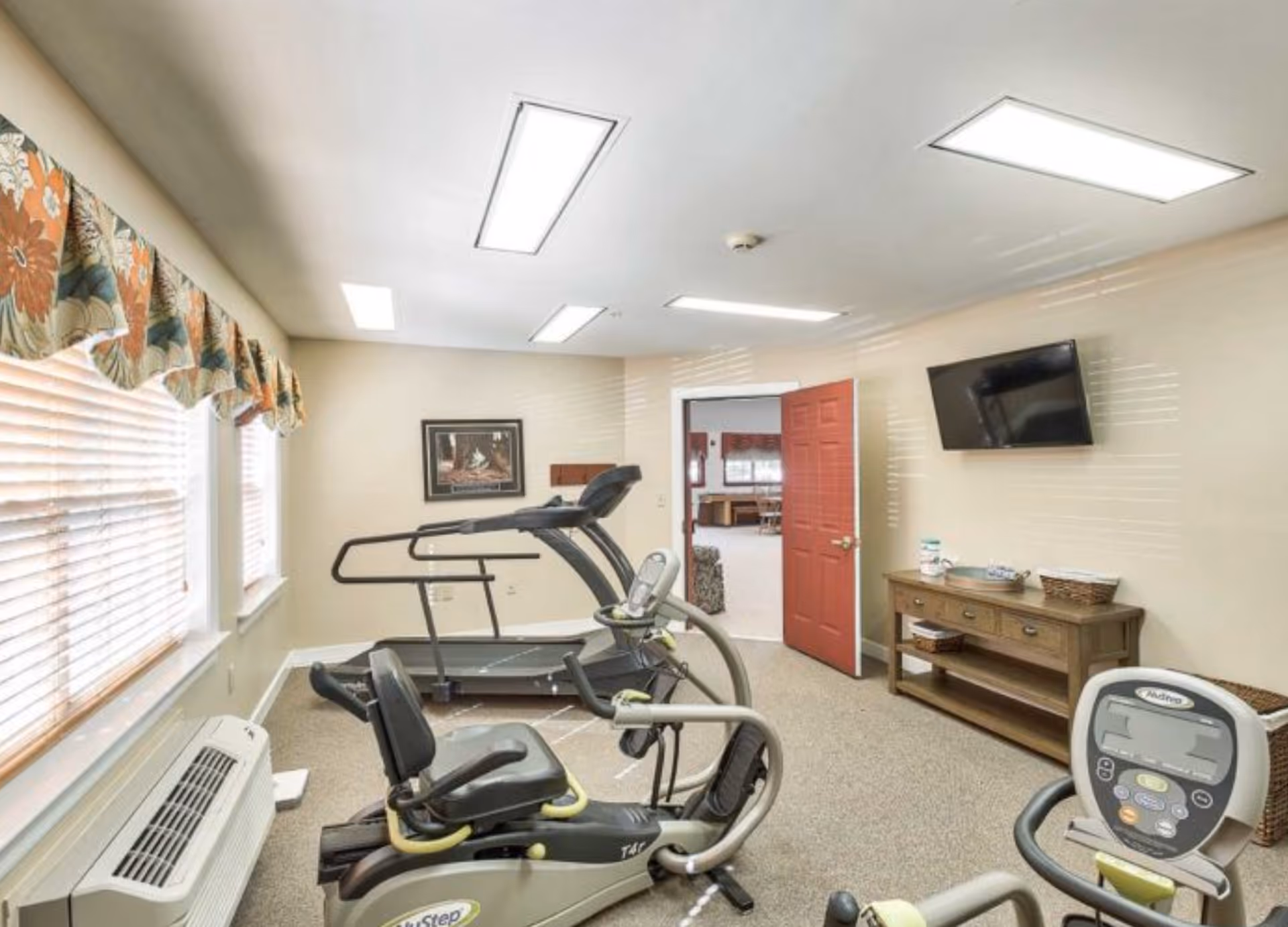 A small exercise room with a treadmill and two stationary bikes facing a wall-mounted TV. The room has beige walls, carpeted floor, and a window with floral valance curtains. A wooden console table with baskets and decorative items is against one wall, and a red door is open leading to another room.