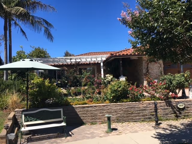 Front exterior view of Ocean Breeze Care Home II showing a garden with various plants and flowers, a bench with a green umbrella, a stone wall, and a clear blue sky with palm trees in the background.