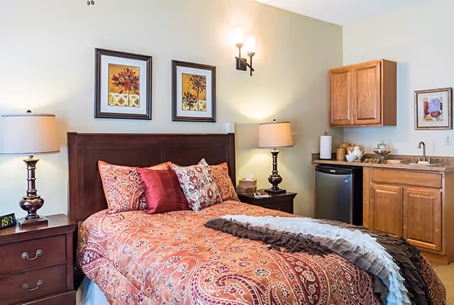 A cozy bedroom with a wooden headboard bed covered in a red and orange patterned bedspread and multiple pillows. There are two framed floral artworks on the wall above the bed. On either side of the bed are dark wooden nightstands with lamps. To the right, there is a small kitchenette area with wooden cabinets, a mini fridge, a sink, and countertop items including a paper towel holder and a glass jar.