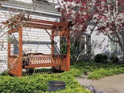 A wooden swing bench under a pergola in a garden area with green shrubs and a tree with red leaves, situated next to a light-colored building with windows.