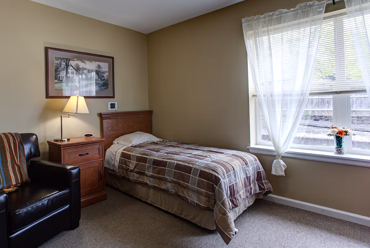 A small bedroom with a single bed covered in a brown plaid comforter, a wooden nightstand with a lamp and alarm clock, a black leather armchair with a striped blanket, a framed picture on the wall, and a window with sheer white curtains and a vase of flowers on the windowsill.