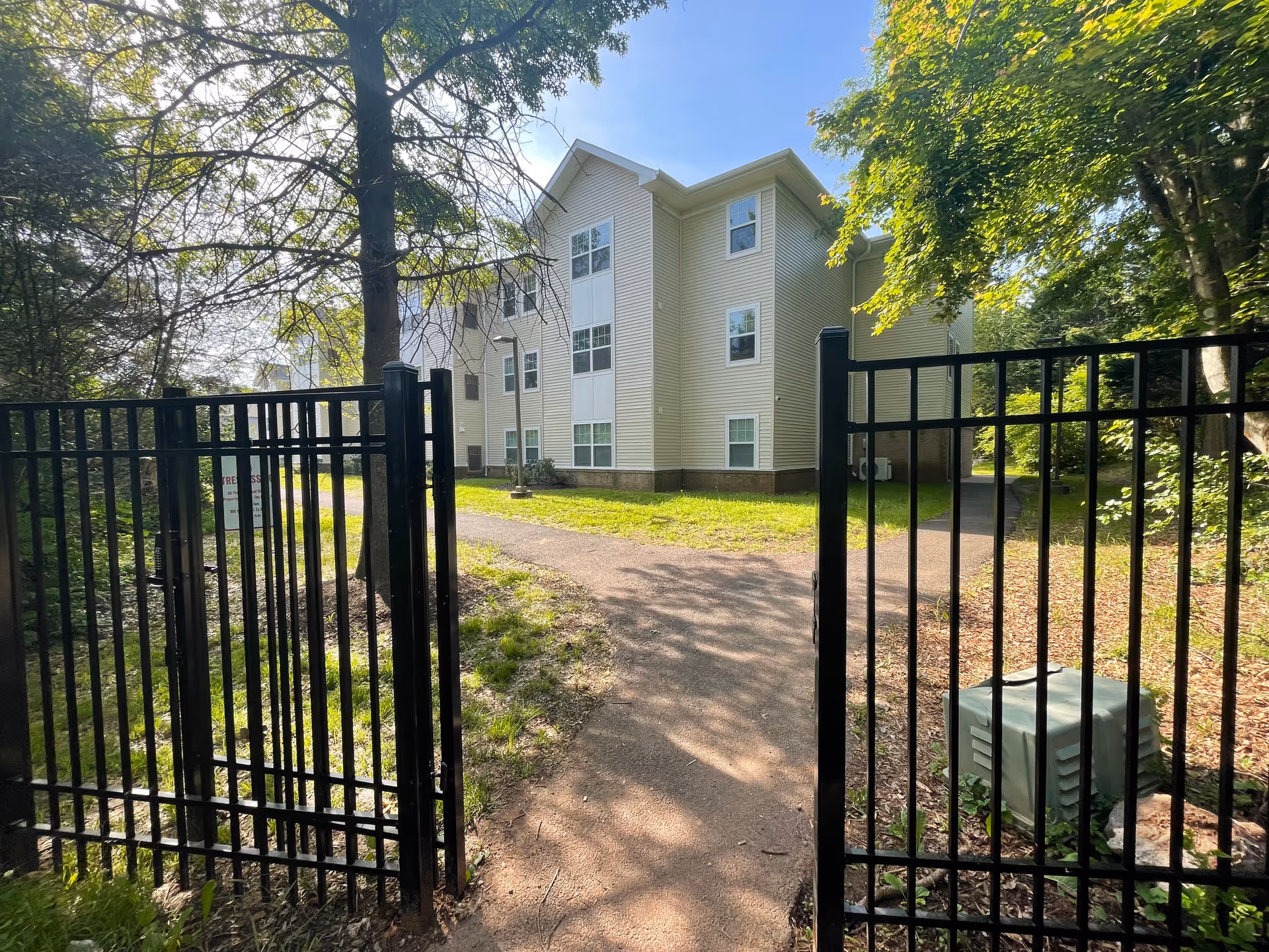 View through a black metal gate of a light-colored three-story apartment building with a paved path and surrounding trees.