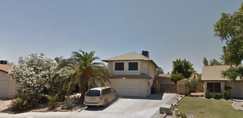A two-story residential building with a garage and a driveway where a minivan is parked. The house is surrounded by trees and shrubs, including a palm tree and flowering bushes. The sky is clear and blue.