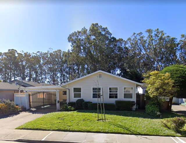 Single-story white house with three windows on the front, a small lawn with a young tree, bushes along the house, and a gated driveway on the left side. Tall trees are visible in the background under a clear blue sky.