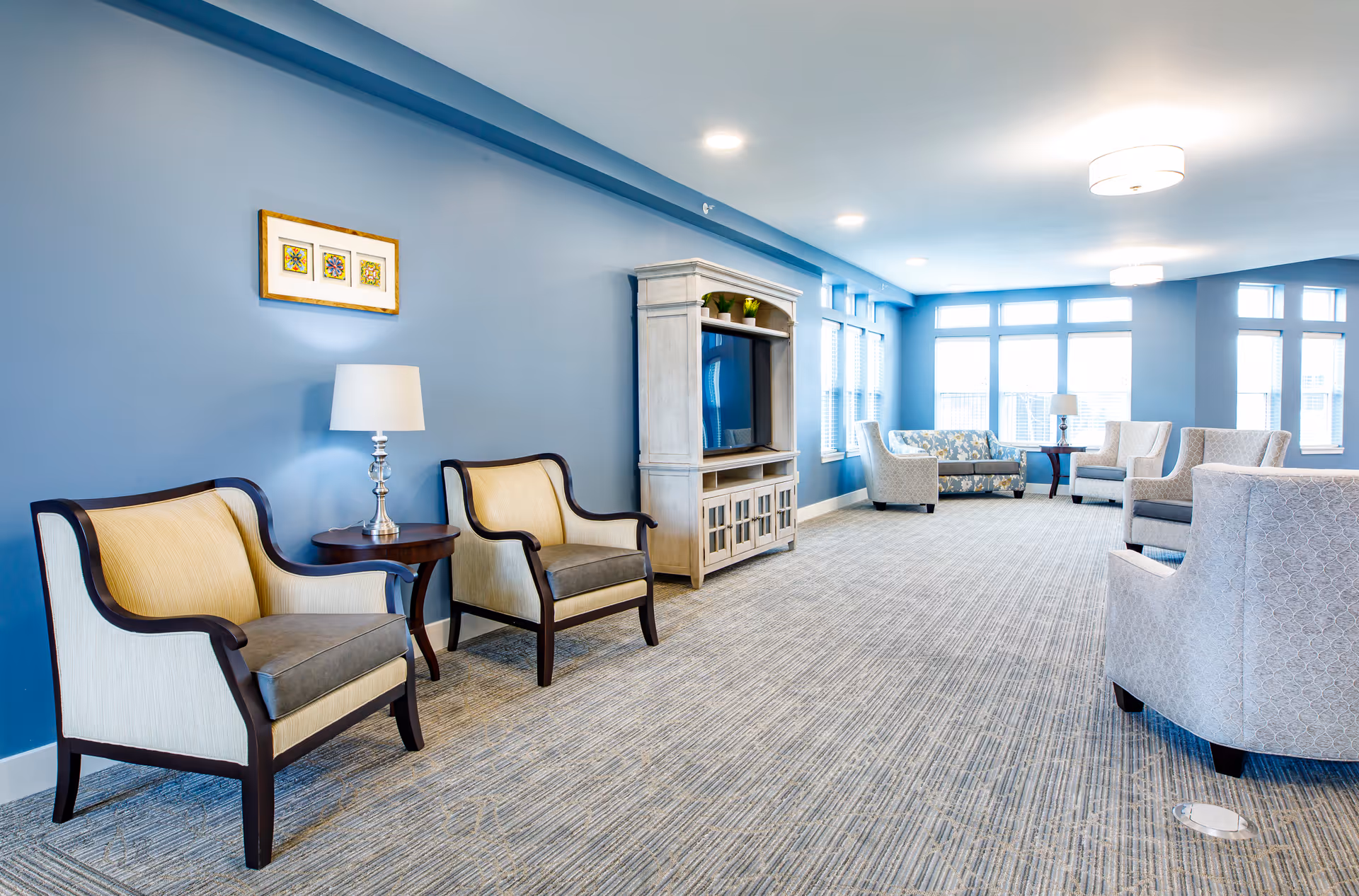 A bright, spacious lounge with blue walls featuring armchairs, side tables, a TV cabinet, and large windows.