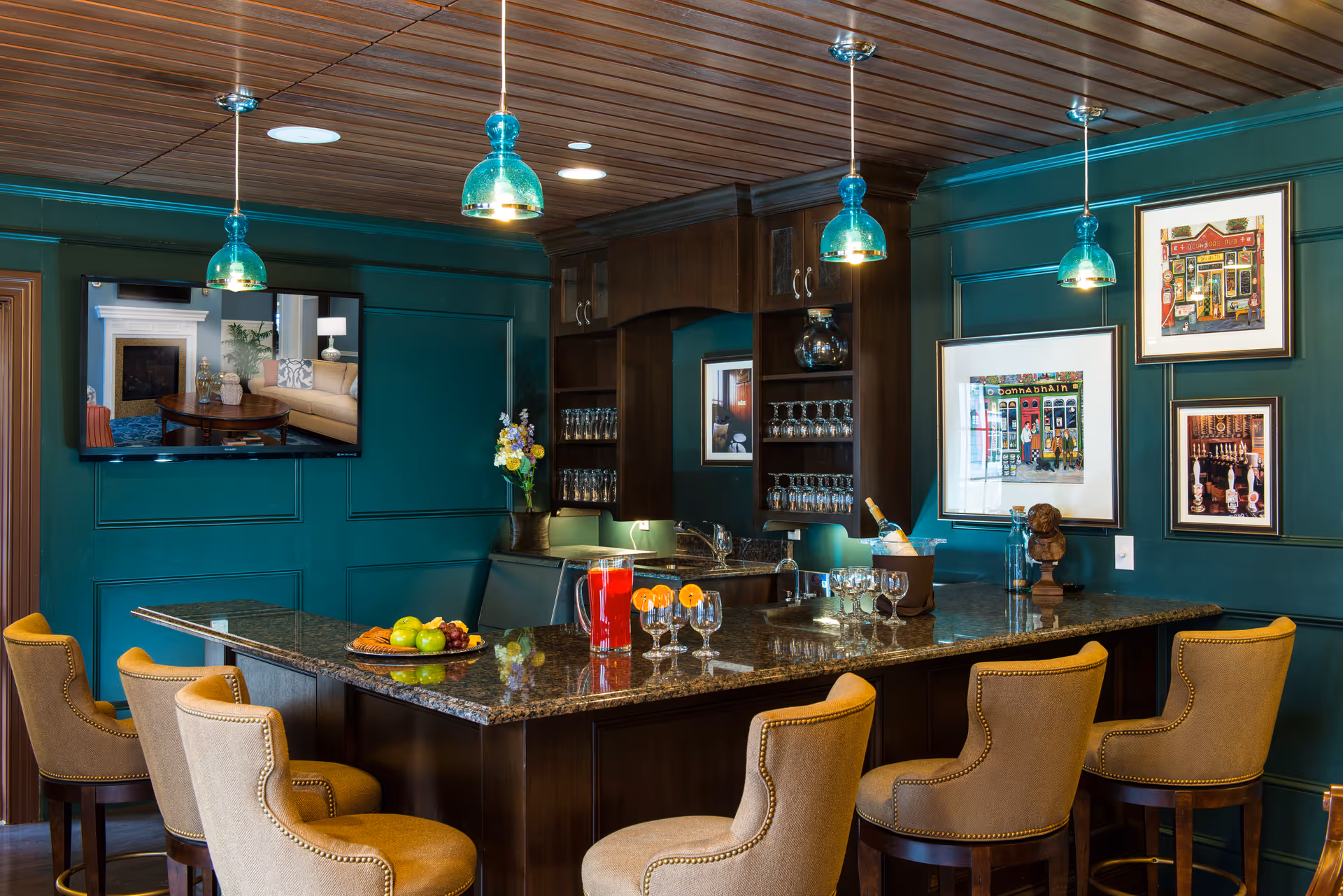 A stylish indoor bar area with a dark granite countertop surrounded by six beige upholstered bar stools. The walls are painted teal and decorated with framed artwork. Above the bar hang four blue pendant lights from a wooden ceiling. The bar features glass shelves with glassware, a sink, a pitcher of red beverage, and several glasses with orange slices on the rim. A flat-screen TV is mounted on the wall showing a cozy living room scene.