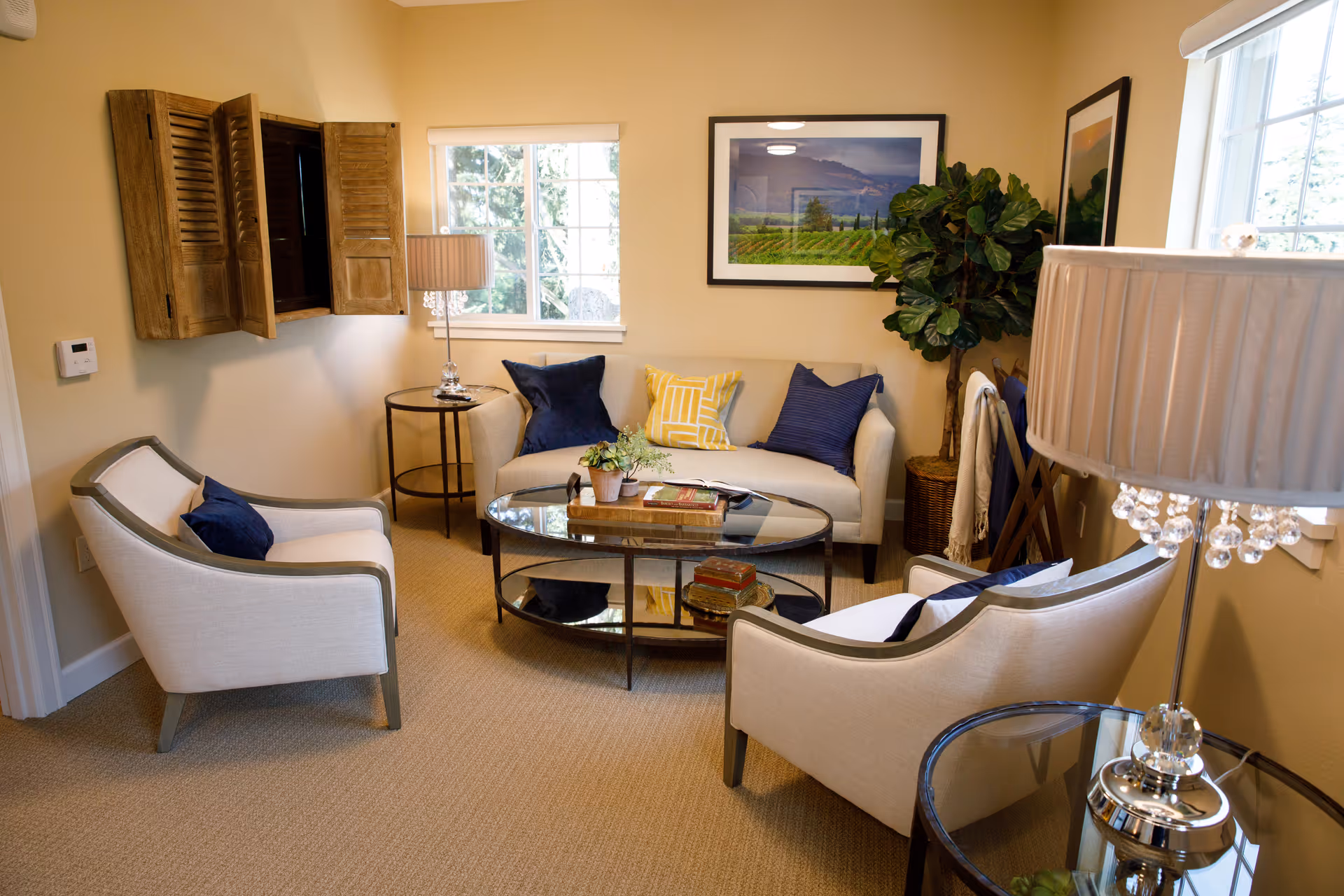 A cozy living room with a beige sofa adorned with blue and yellow pillows, two matching armchairs with blue pillows, a round glass coffee table with books and a small plant, a side table with a lamp, framed landscape artwork on the walls, and two windows letting in natural light.