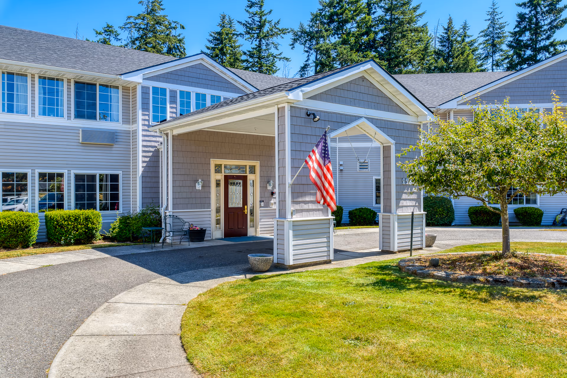 Exterior view of Laurel Place Assisted Living & Memory Care building entrance with a covered porch, American flag, well-maintained lawn, and trees under a clear blue sky.