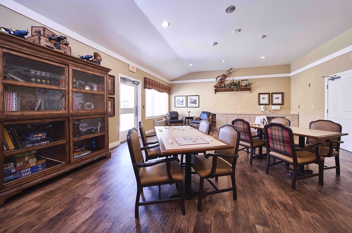 A well-lit common room with wooden flooring, featuring two tables with chairs arranged around them. One table has a Scrabble game set up. There is a large wooden cabinet filled with books and board games on the left side. The walls are decorated with framed pictures and a shelf holding a violin and plants. Two armchairs are positioned near a window with a plaid valance.